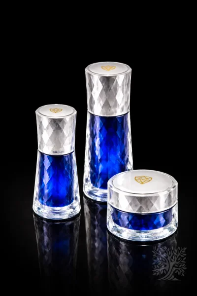 Clear and blue dual-tone faceted acrylic skincare bottles and cosmetic jar set with gold emblem lids on black surface.