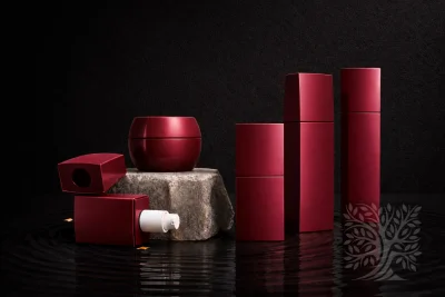 Deep red cosmetic bottles and matching jars with smooth finishes, arranged on a stone base with soft reflections.