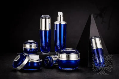 Deep cobalt blue acrylic skincare bottles and cosmetic jars with chrome accents, arranged on black blocks with open jar detail.