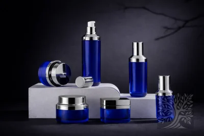 Cobalt blue acrylic skincare bottles and cosmetic jars with silver caps, arranged on white blocks with pump and jar display.